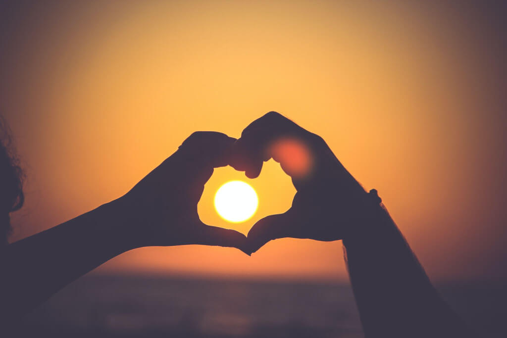 An image of a person using their two hands to form the shape of a heart with a sunset in the middle