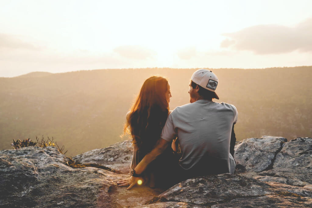 A couple sitting on the ridge of a hill looking out into the sunset.